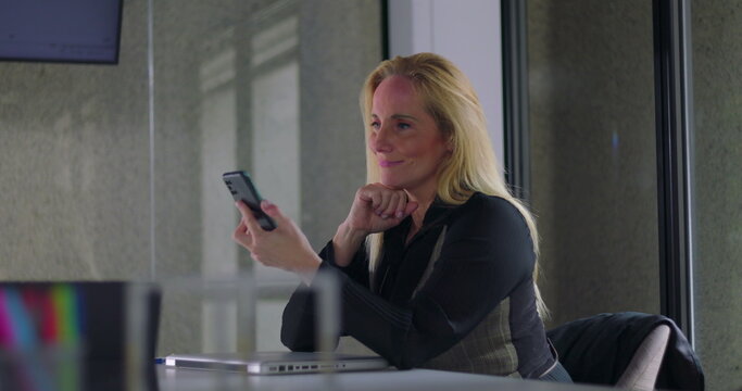 Business woman smiling while using smartphone at desk during work day in modern office setting, engaged in communication or checking messages - Powered by Adobe