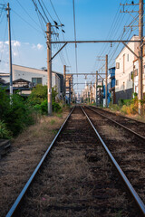 Fototapeta premium Ground level view shows narrow gauge tracks through an Osaka neighborhood, with utility poles, catenary wires, and houses. Late afternoon light casts long shadows.