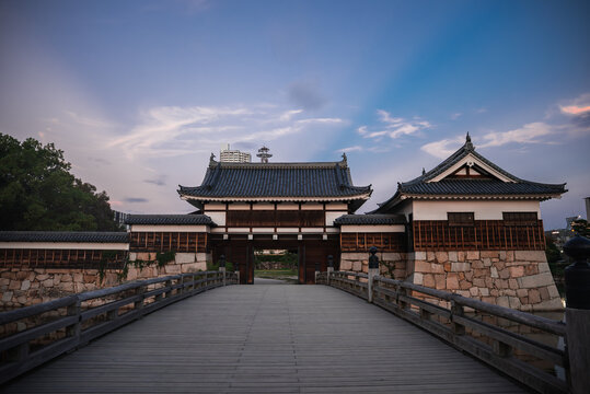 A wide wooden bridge leads to a gatehouse at Osaka Castle in Osaka, Japan at dusk. Tiled roofs and white walls stand centered as modern buildings rise beyond trees.