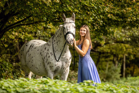 Young woman in a flowing blue dress posing beside her Danish Knabstrupper horse in a peaceful green natural setting outdoors.