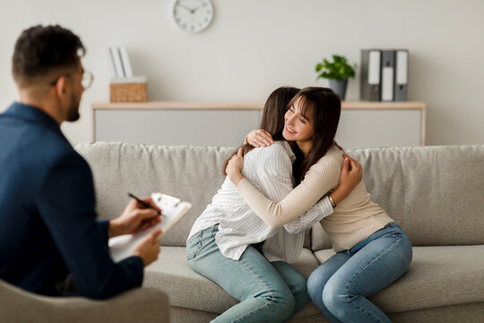 A male psychologist sits with a notepad while two Arab women embrace on a sofa in a cozy office. They are finding solutions to their relationship issues through therapy.