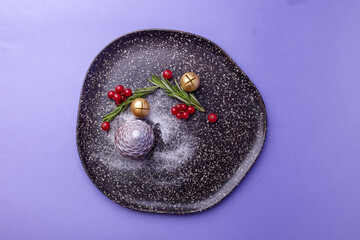 Festive dessert on dark speckled plate with rosemary, red berries and gold ornaments against purple