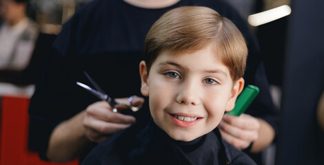 Smiling caucasian young boy at hair salon with hairdresser and scissors, retro barbershop banner