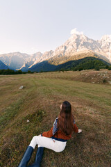 Fototapeta premium Woman lying on meadow looking at towering mountains and serene nature landscape, hiking travel scene in wide open outdoors field with boots, long hair and peaceful sky.