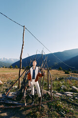 Fototapeta premium Man in traditional clothing stands by a rustic fence in a rural mountain landscape and grassy field under clear sky. Outdoor portrait, countryside lifestyle, scenic nature scene.