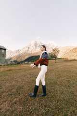Fototapeta premium Woman walking in countryside meadow wearing a knit sweater and boots, scenic mountains in background, autumn light, rural cottage nearby, outdoor lifestyle portrait and travel vibe