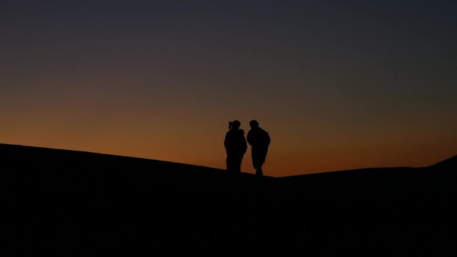 Two Berber men walking in silhouette during a warm Sahara Desert sunset.