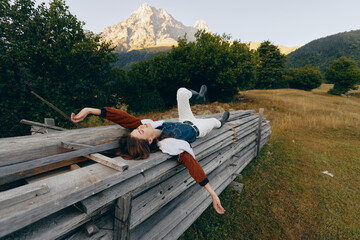 Fototapeta premium Woman lying relaxed on wooden fence in meadow with mountain backdrop, portrait of outdoors leisure and nature escape, casual clothing and serene summer landscape.