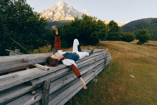 Woman on wooden fence in mountain meadow, relax outdoors among trees and nature with distant peak, casual outfit and carefree pose for peaceful escape and leisure.