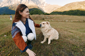 Fototapeta premium Woman smiling and petting a dog in a meadow by the mountains, outdoor nature portrait showing countryside companionship, joyful bonding and relaxed weekend adventure in grassy field.