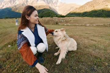 Fototapeta premium Woman crouching in a mountain field smiling while petting a dog, enjoying nature and companionship in a scenic outdoor landscape, casual jacket, happy moment with her pet