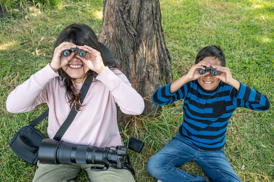 Mother and son relaxing by a tree, smiling while observing nature with binoculars