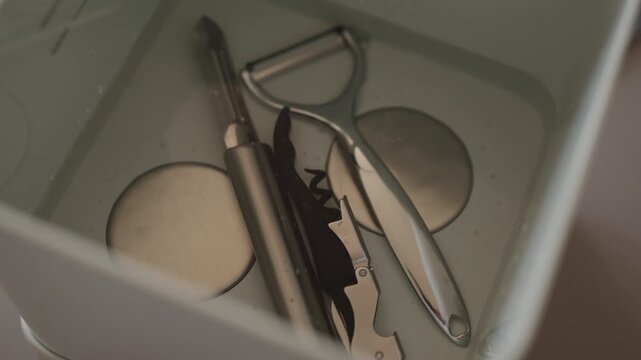Kitchenware receiving deep ultrasonic cleaning, immersing peeler, corkscrew and other metal utensils in water bath, ensuring hygiene and sanitation for home cooking, close-up, nobody, slow motion.