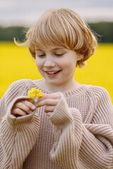 Caucasian girl, 9 years old, blonde with short hair, wearing beige knitted sweater, front view, smiling and holding yellow flower in field with forest background. Concept childhood, joy, nature.