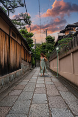 A narrow stone alley in Kyoto is lined by wooden machiya and a plaster wall with lantern. A woman walks away at dusk, warm low light and pink clouds above.
