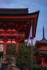 Obraz premium Vermilion temple buildings and pagoda rise at Kiyomizu dera in Kyoto, Japan. A Japanese flag and stone guardian statues stand in the foreground in early evening light.