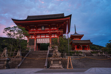 Naklejka premium A vermilion gate, pagoda, and Niomon gate stand at Kiyomizu dera in Kyoto at dusk. Lion dog statues, stone stairs, and latticework appear against forested hills.