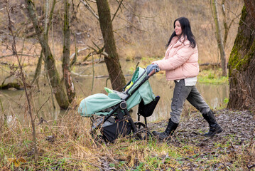 A mother walks with her child in a stroller in the park along the river in late autumn.