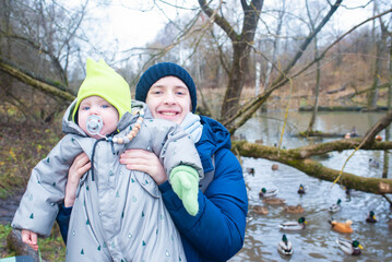 A teenage boy holds his baby brother. The boys stand in front of a river with ducks in a park.