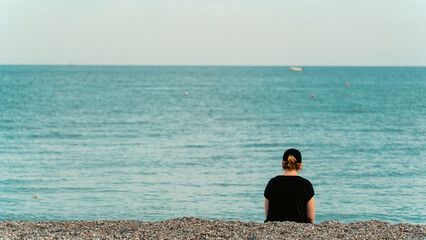 A woman seen from behind, looking at the sea from a pebble beach, deep in thought. Reflective and serene mood, perfect for lifestyle and emotional concepts.
