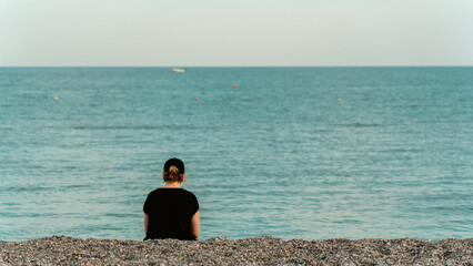 A woman seen from behind, looking at the sea from a pebble beach, deep in thought. Reflective and serene mood, perfect for lifestyle and emotional concepts.