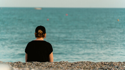 A woman seen from behind, looking at the sea from a pebble beach, deep in thought. Reflective and serene mood, perfect for lifestyle and emotional concepts.