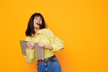 A joyful young woman dances with a tablet in her hands, wearing a yellow shirt and jeans, against a vibrant orange background, conveying energy, creativity, and playful work moments.