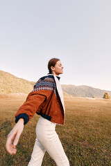 Fototapeta premium Woman walking in countryside field wearing patterned jacket and cozy sweater, enjoying autumn outdoors near mountains with sunlight, relaxed casual style and free travel lifestyle.