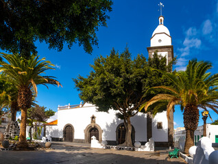 A stunning view of the historic church in Arrecife, Lanzarote, surrounded by tall palm trees. The bright blue sky enhances the picturesque scene, capturing the essence of this charming location