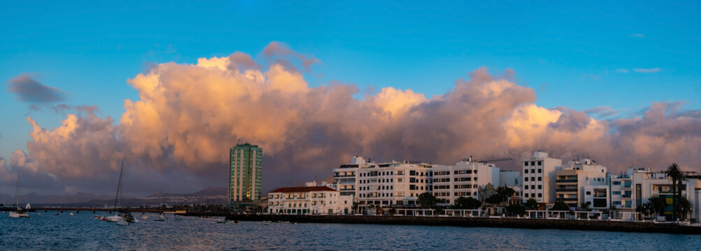 Arrecife Lanzarote waterfront view featuring historical buildings and people leisurely walking along the promenade. The serene atmosphere and clear sky create a picturesque scene on sunrise