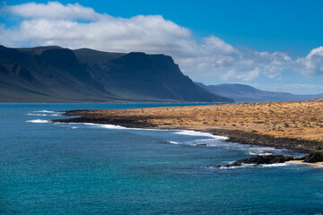 Rugged volcanic coastline of La Graciosa features turquoise waters meeting black rocks. Majestic cliffs rise in the distance under blue skies. Visitors walk along the sandy path
