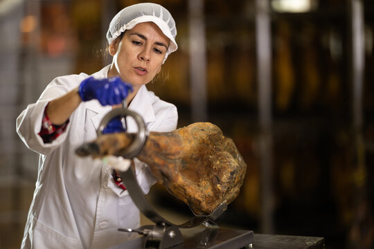 Qualified female specialist works in a meat factory, she has put the ham on a stand and is preparing to slice it. Process of making jamon