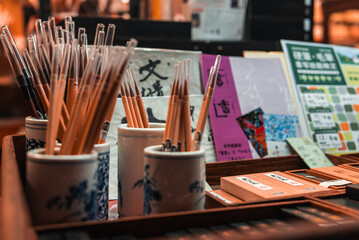 Ceramic cups hold wooden pencils and clear capped pens beside notebooks with Japanese text in a Nara shop, warm indoor light and shallow depth of field emphasize detail.
