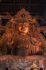Obraz premium A large gilt Buddhist deity stands in a dim wooden hall in Nara, Japan, with a raised hand, halo like rays, crown, and layered robes lit by warm daylight.