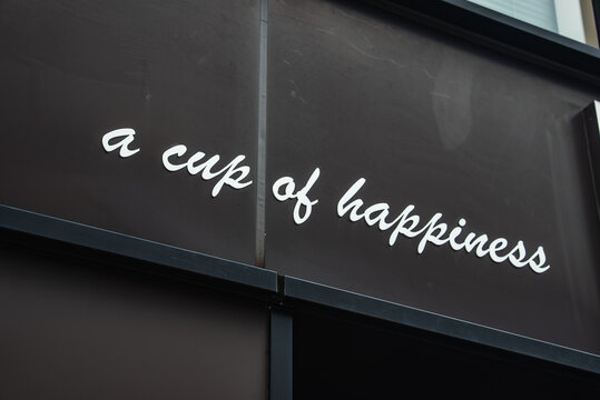 White cursive text spells a cup of happiness on a dark matte facade. Soft daylight and a tight angled view emphasize clean typography and a modern urban mood.