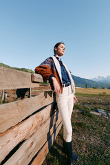 Fototapeta premium Woman standing and leaning on wooden fence in countryside field with mountains background, portrait style showing sweater, boots and casual fashion in scenic outdoor landscape.