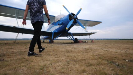 Pilot approaches vintage blue biplane on grassy airstrip, showcasing the anticipation of flight as he walks confidently towards the aircraft, ready for takeoff and adventure in the sky