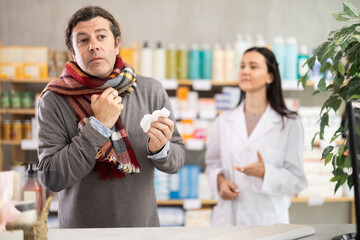 Man in scarf with sore throat is looking for medicine in a pharmacy. Female pharmacist is helping him