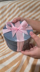 Hands holding a round, textured blue-gray gift box tied with a bright pink satin ribbon bow; elegant present for a birthday, anniversary, or holiday celebration on striped fabric.