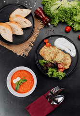 Tomato soup and zucchini fritters with yogurt sauce, salad and bread on dark table