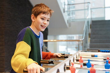 Smiling boy in colorful sweater happily playing foosball table game indoors. Happy childhood.