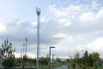 Stadium-style floodlight mast with ladder rises above trees and walkway in a city park. LED array used for park illumination under partly cloudy sky, daytime perspective.
