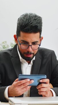 Indian businessman playing on smartphone mobile application celebrate winning racing or shooter game at office desk workplace. Professional manager freelancer man enjoy testing good work of new app