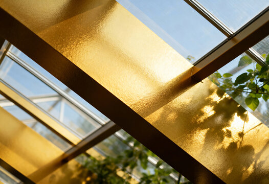 Close-up of a textured gold beam in a modern glass structure. Sunlight and shadows from plants create an abstract architectural background - Powered by Adobe