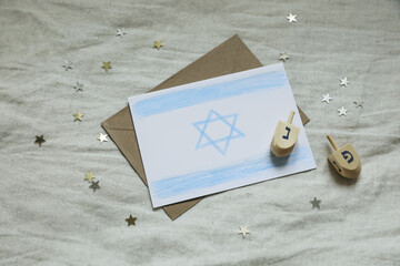 Hanukkah holiday flat lay top view. Blank greeting card, envelope. Hand drawn Israeli flag. Wooden dreidel spin toys, golden star confetti. Fabric background. Jewish winter celebration mockup template