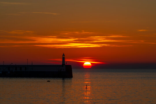 Dramatic orange sunset over a harbour breakwater and lighthouse silhouette in Peel, the Isle of Man.