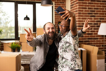 Excited multiethnic couple stand surrounded by moving boxes, taking break to capture selfie with cellphone. White bearded man makes peace sign as girlfriend holds mobile device to snap photo.