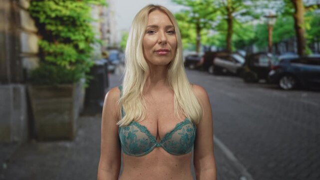 Woman with blonde hair wearing green lace bra on city street looks sideways against parked cars; confidence.