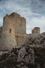 Close-up view of the medieval fortress of Rocca Calascio in Abruzzo, perched on a rocky mountain ridge. Ancient stone walls and towers stand against a dramatic cloudy sky with the mountains.