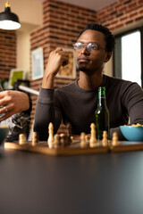 Portrait of pensive black man sitting at table with chessboard and beer in front of him. African american guy wearing glasses, contemplating his next move during friendly chess match with friends.
